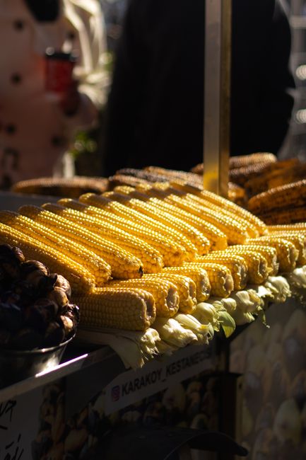 A close-up view of a street food stall displaying freshly grilled corn on the cob, lined up in neat rows with golden-yellow kernels and some slightly charred pieces at the edges. The corn is arranged